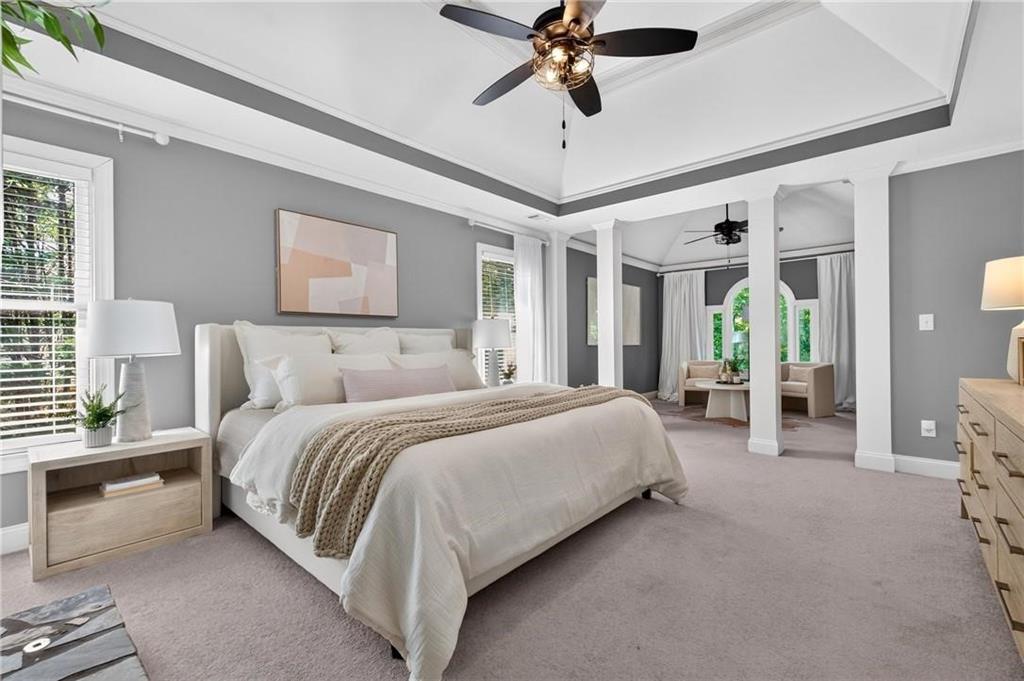 Staged primary bedroom with a white bed, tray ceiling, and adjoining sitting area.