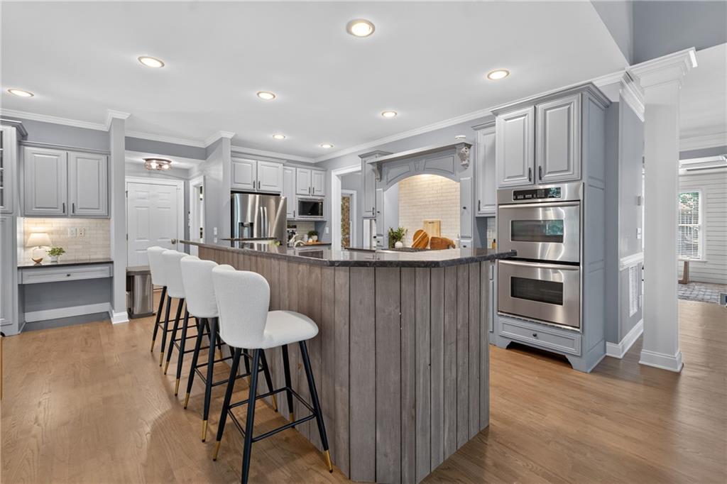 Kitchen with a large wood-paneled island, white bar stools, gray cabinetry, and stainless steel double ovens.