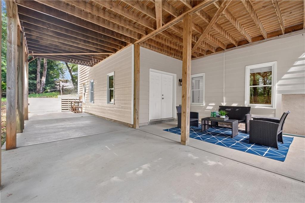 Covered patio with exposed timber beams, concrete floor, and wicker seating on a blue rug.