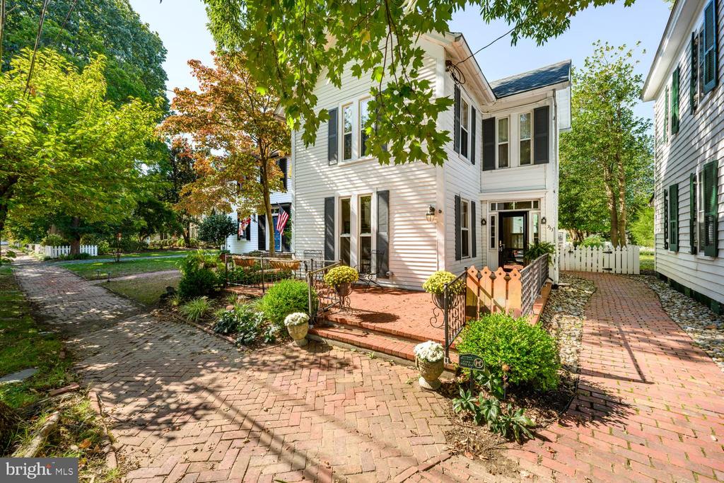 White two-story house with dark shutters, brick walkways, and leafy trees around the entrance.