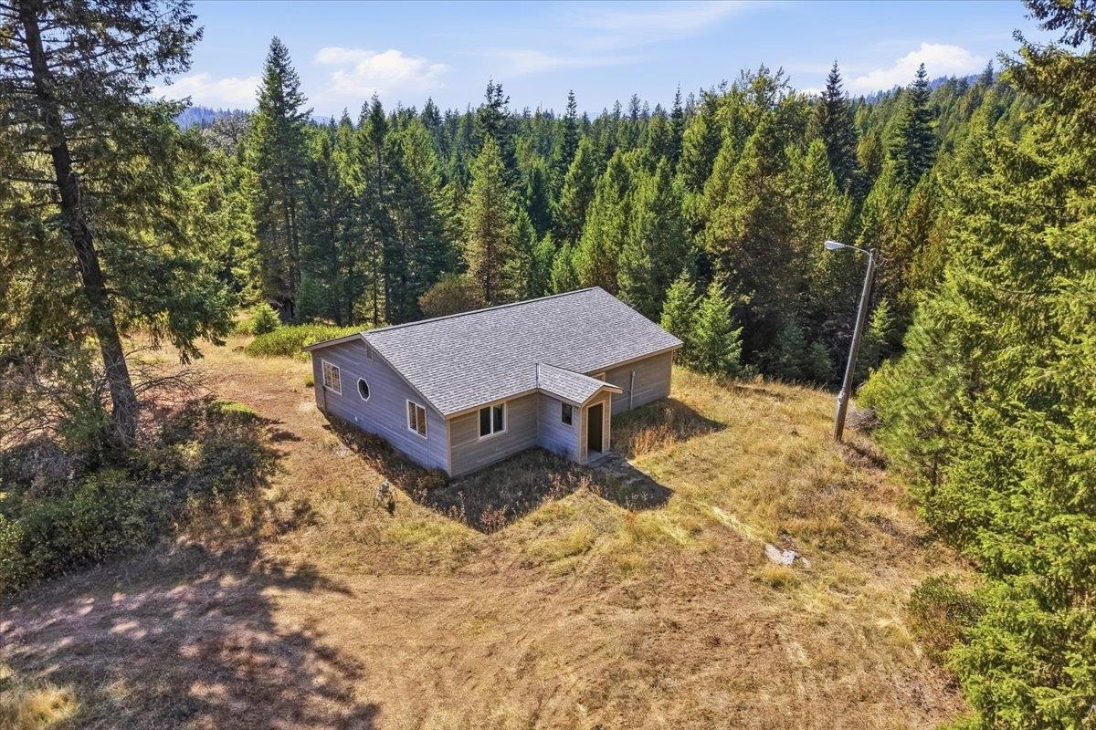A gray single-story house sits in a grassy clearing surrounded by evergreen forest.