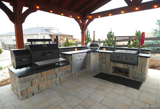 Outdoor kitchen beneath a wooden pergola, featuring a stone island, built-in grill, and sink.