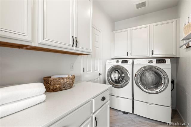 Laundry room with white front-loading washer and dryer beneath cabinets beside a counter with folded towels.