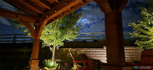 Covered patio with a wooden pergola, seating, illuminated trees, and a stone wall at night.