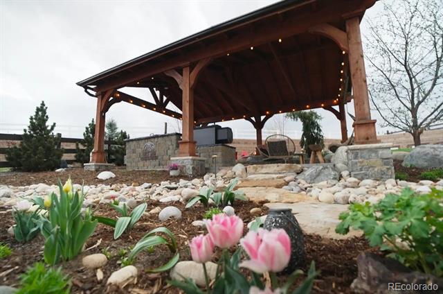 Covered wooden pavilion with string lights above a stone garden and blooming flowers.