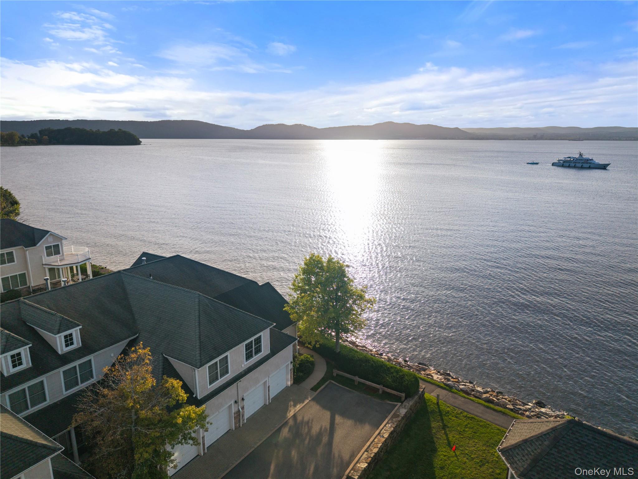 Aerial view of waterfront homes beside a sunlit lake, with a yacht and wooded hills beyond.