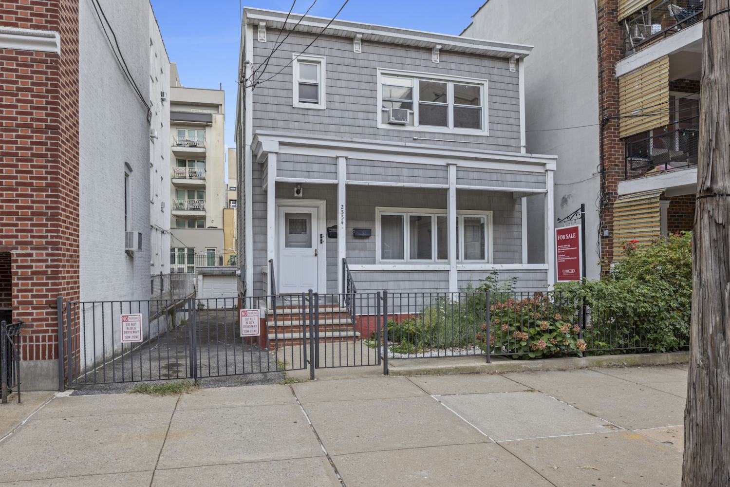 Gray two-story shingle house with a covered porch, fenced garden, and for-sale sign.