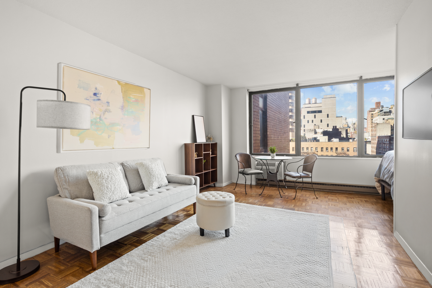 Furnished living room with parquet floors, a gray sofa, and large windows overlooking city buildings.
