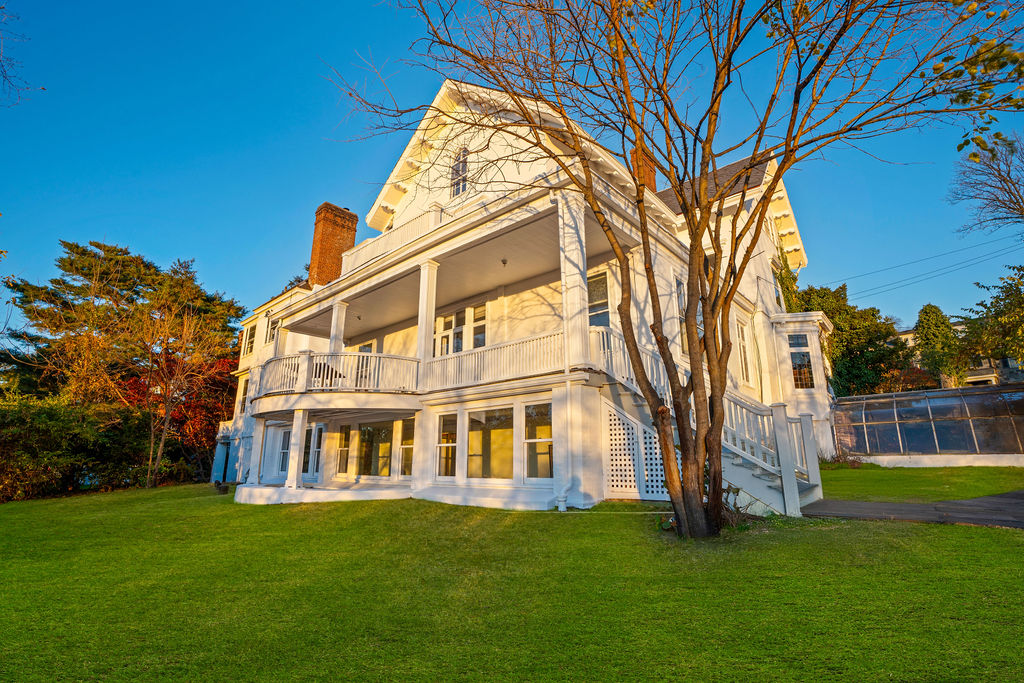 White multi-story house with a covered porch and balcony, partly screened by a leafless tree.