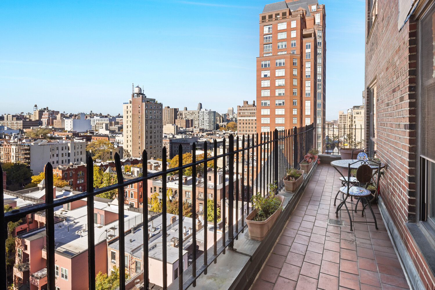 Apartment balcony with potted plants and outdoor seating overlooking city rooftops.