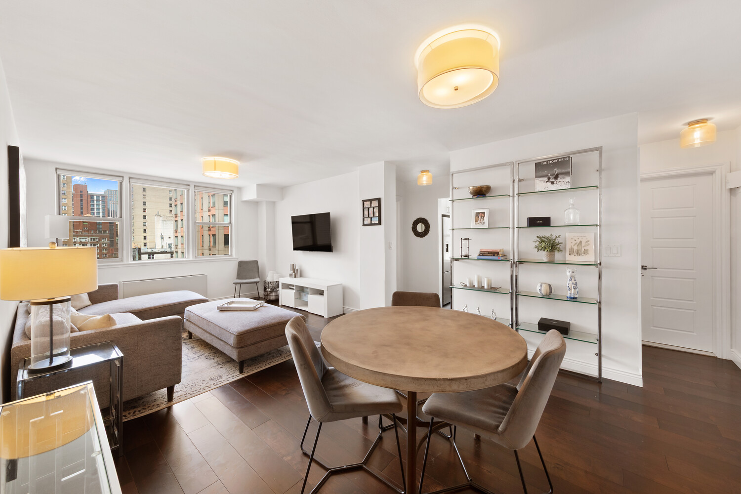 Furnished apartment living and dining room with large windows overlooking city buildings.