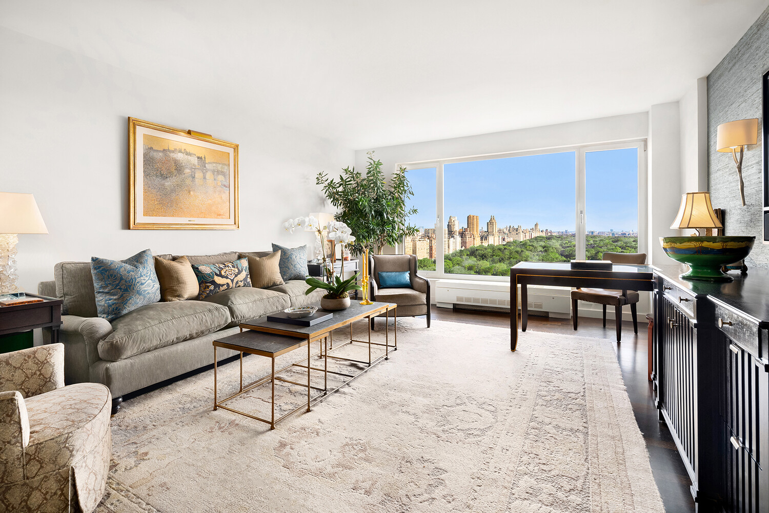 Furnished living room with a gray sofa and wide windows overlooking a green park and city skyline.