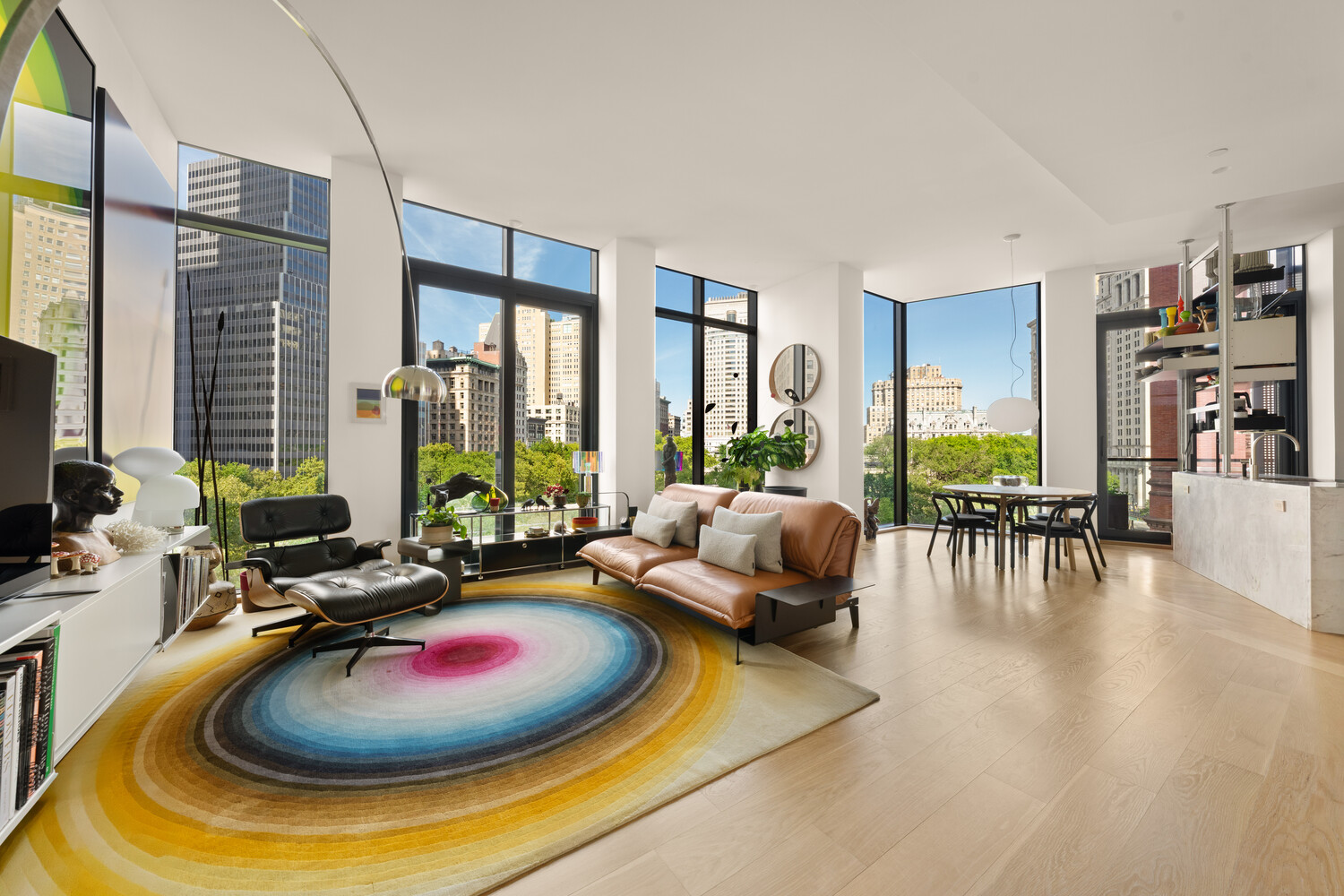 Living room with floor-to-ceiling city-view windows, leather seating, and a colorful circular rug.