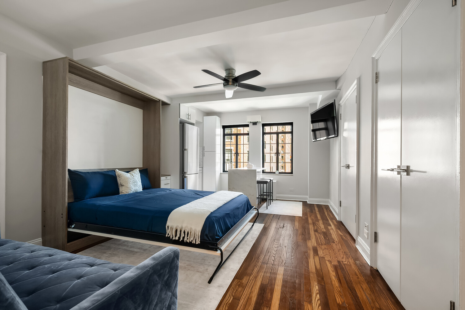 Studio apartment with a blue Murphy bed, matching sofa, hardwood floors, and compact kitchen.