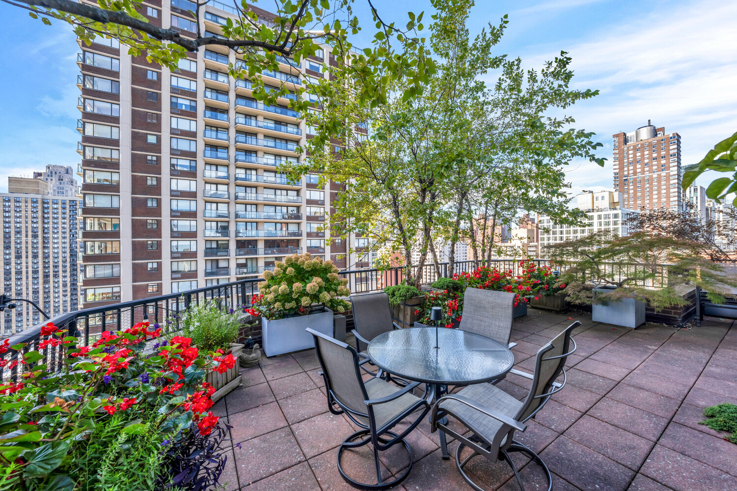 Rooftop terrace with a glass dining table, flowering planters, and views of city apartment buildings.