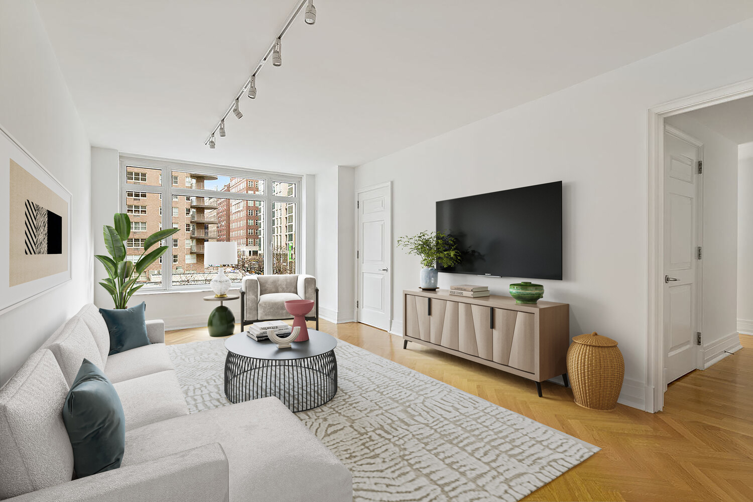 Staged apartment living room with a white sectional, round coffee table, and large window overlooking city buildings.