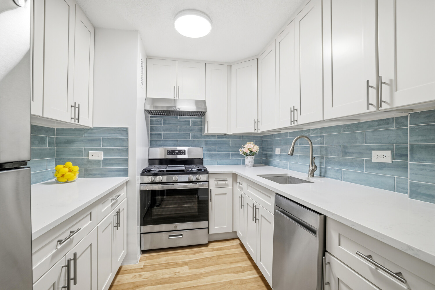 Bright kitchen with white cabinetry, blue tile backsplash, stainless steel appliances, and wood flooring.