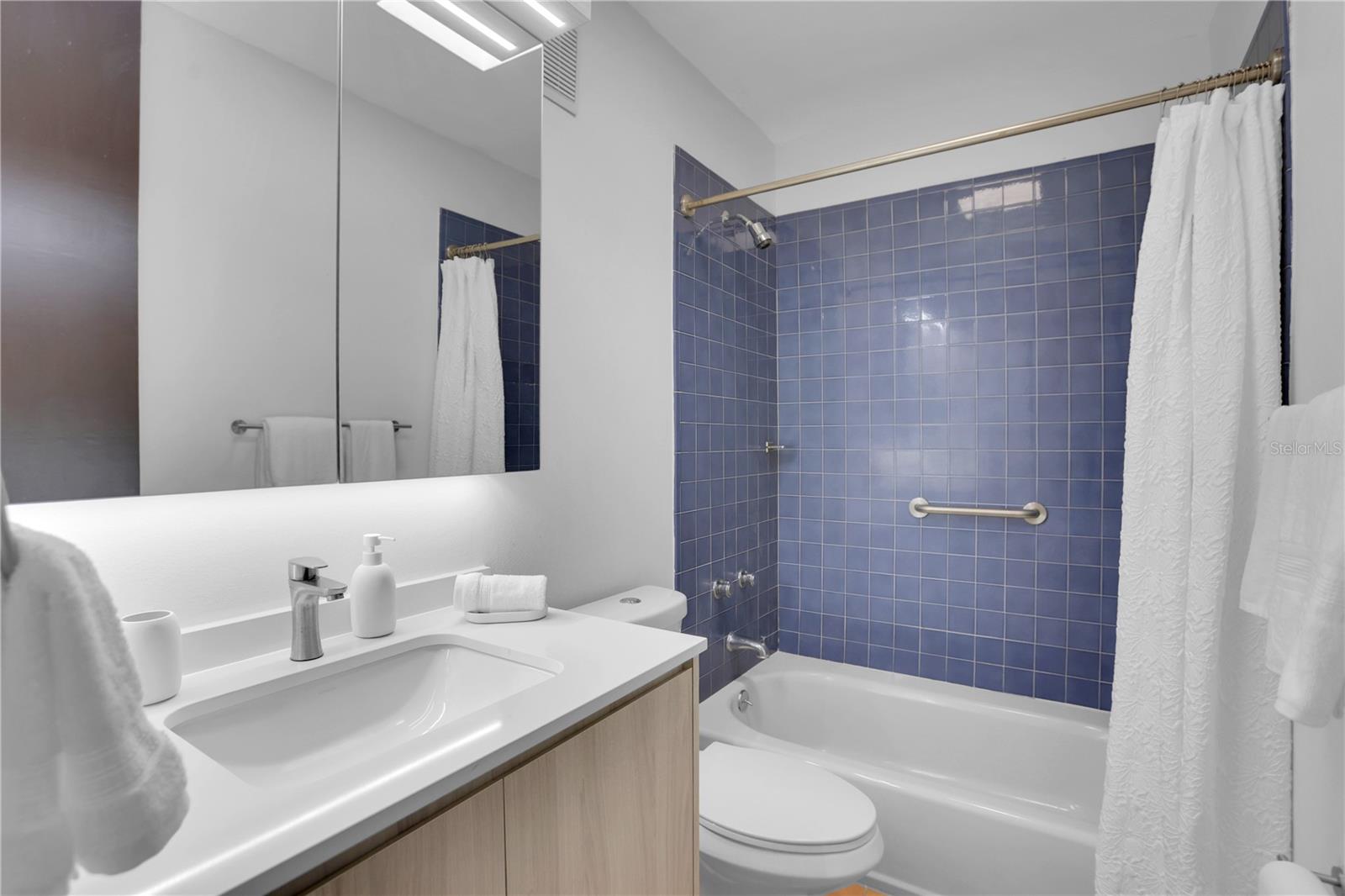 Bathroom with blue tiled tub surround, white vanity, mirrored cabinet, and grab bar.
