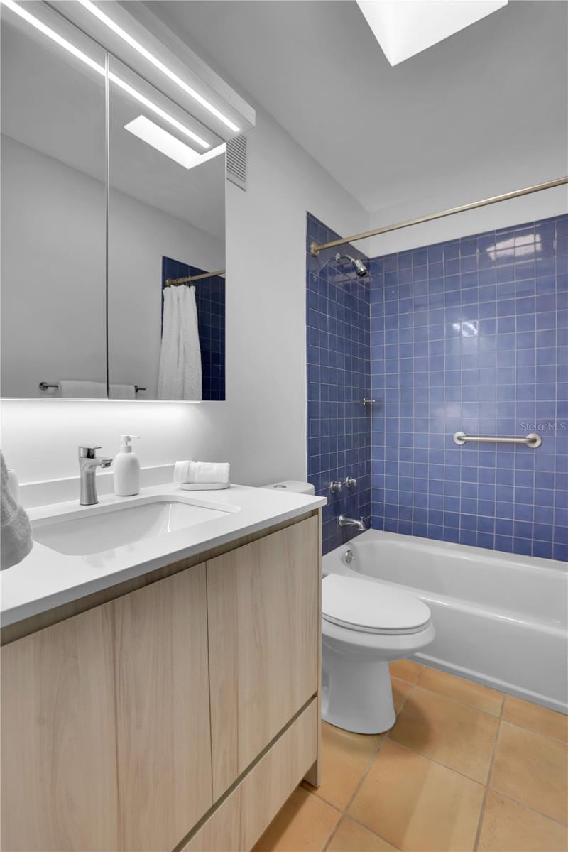 Bathroom with blue-tiled tub and shower, grab bar, and light wood vanity.