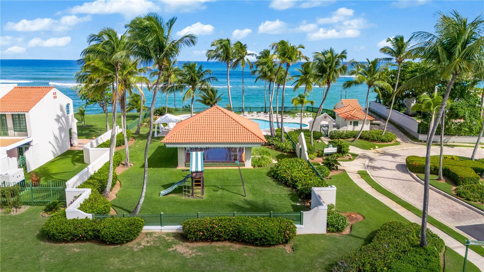 Aerial view of beachfront homes, a pool, and palm-lined lawns beside the ocean.