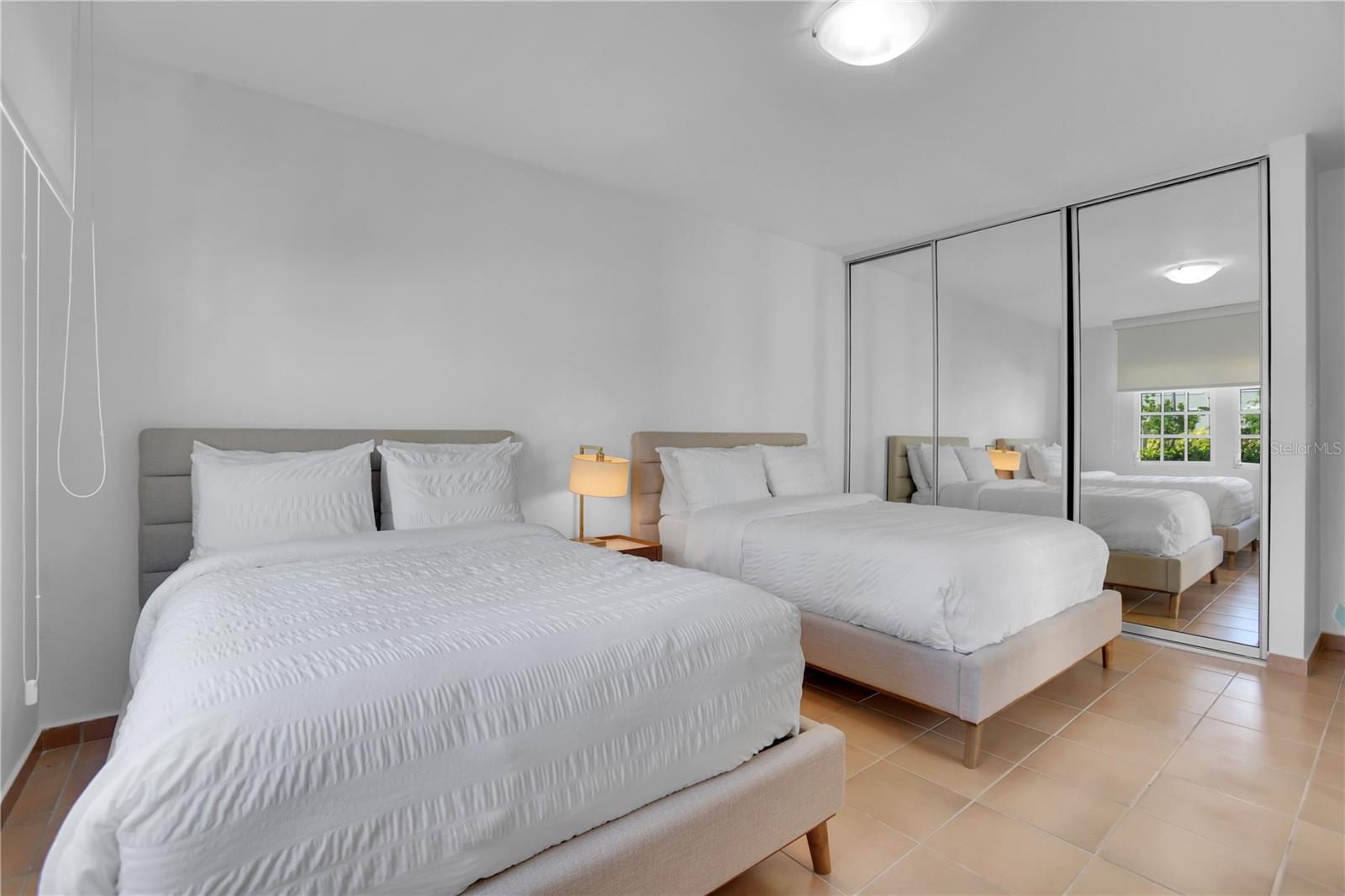 Bedroom with two white beds, mirrored closet doors, and tan tile flooring.