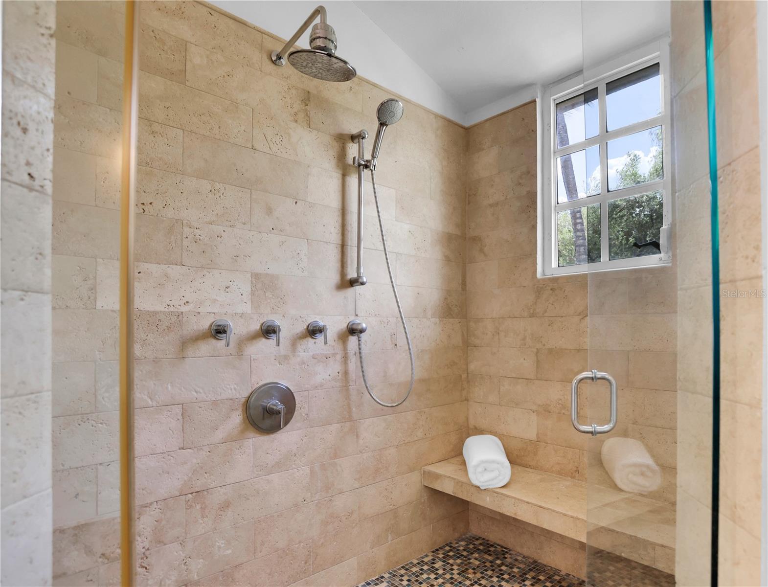 Walk-in shower with beige stone tile, rainfall showerhead, handheld sprayer, and built-in bench beneath a window.