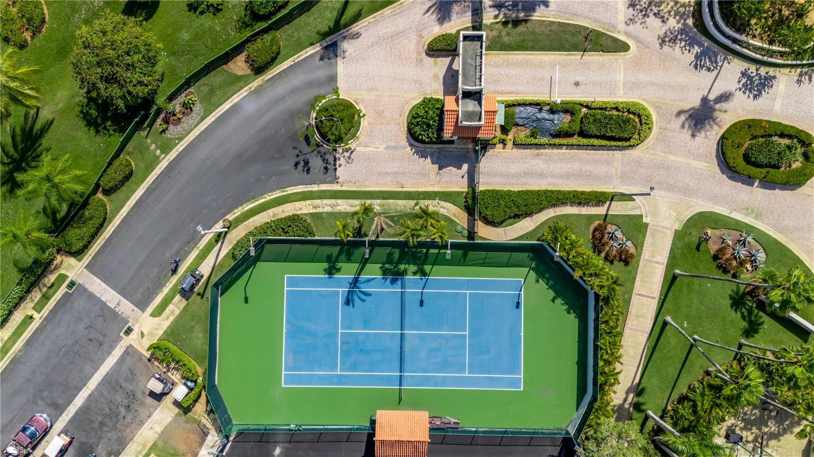 Aerial view of a blue tennis court amid landscaped grounds and paved paths.