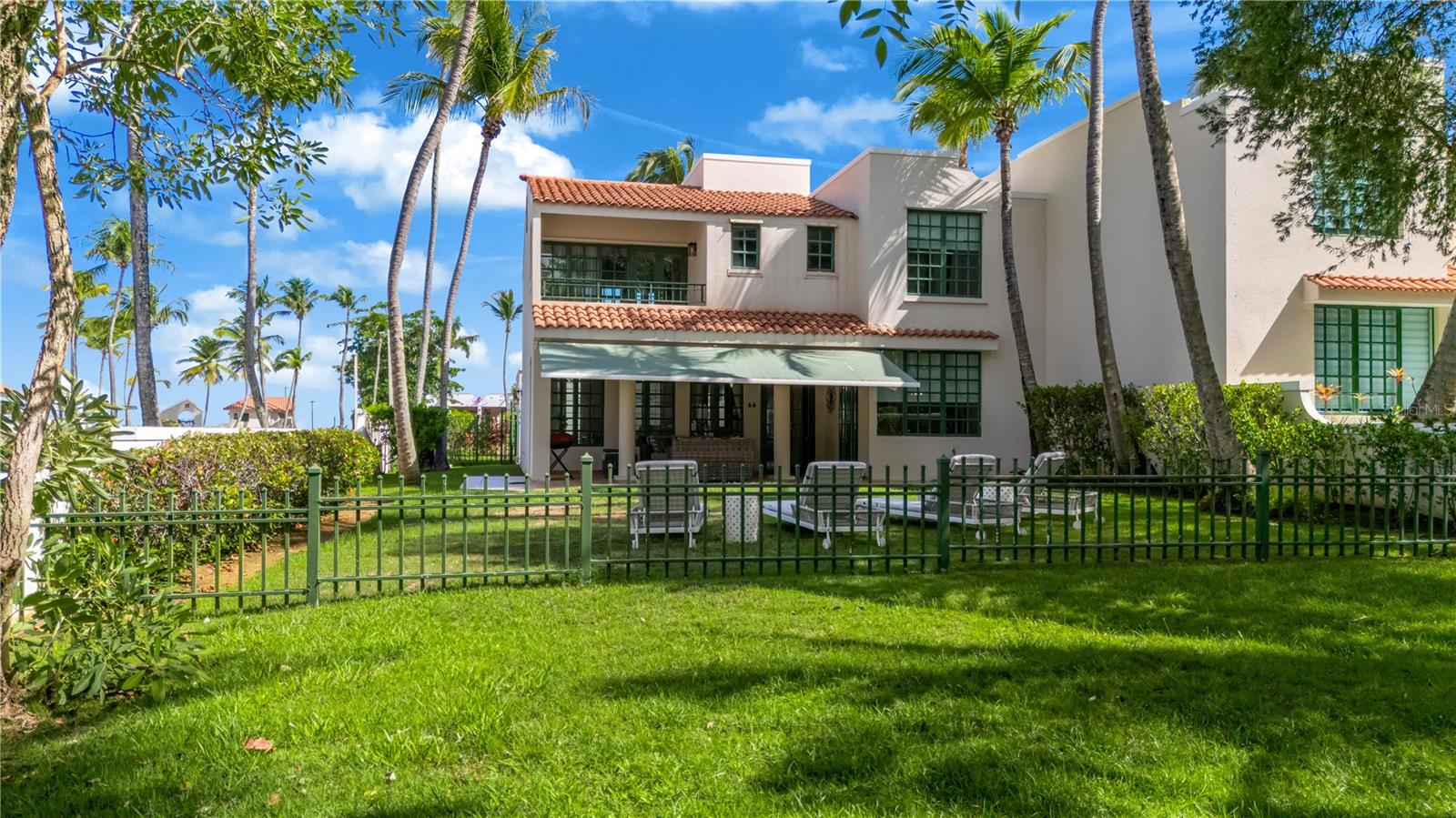Two-story white stucco home with terracotta roofs, a shaded patio, and a palm-lined fenced lawn.