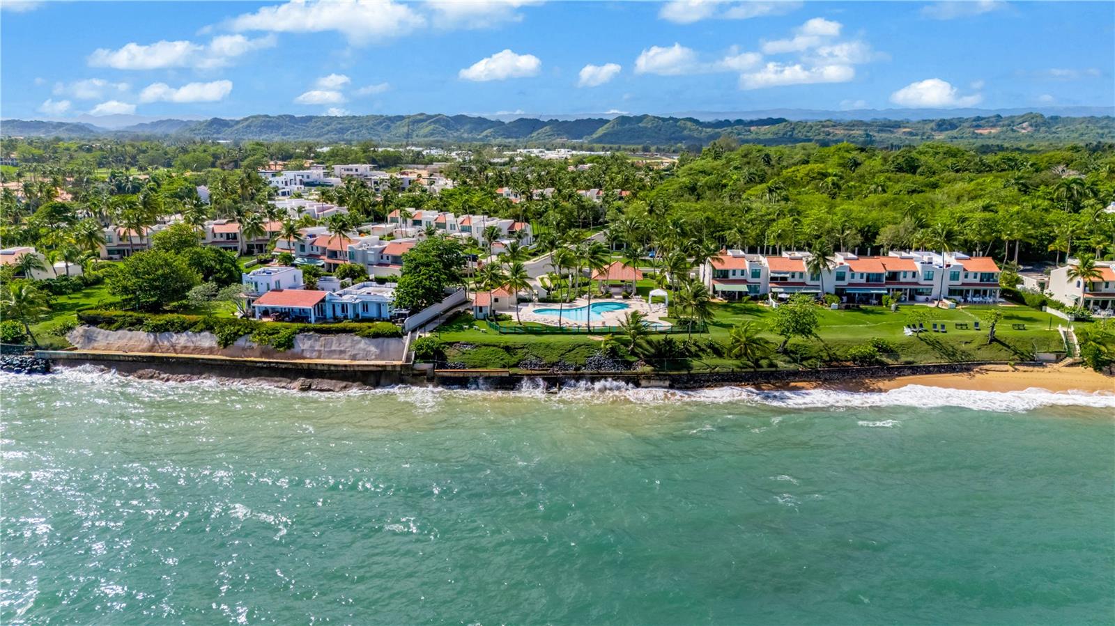 Aerial view of beachfront homes and a pool beside turquoise water, with green mountains in the distance.