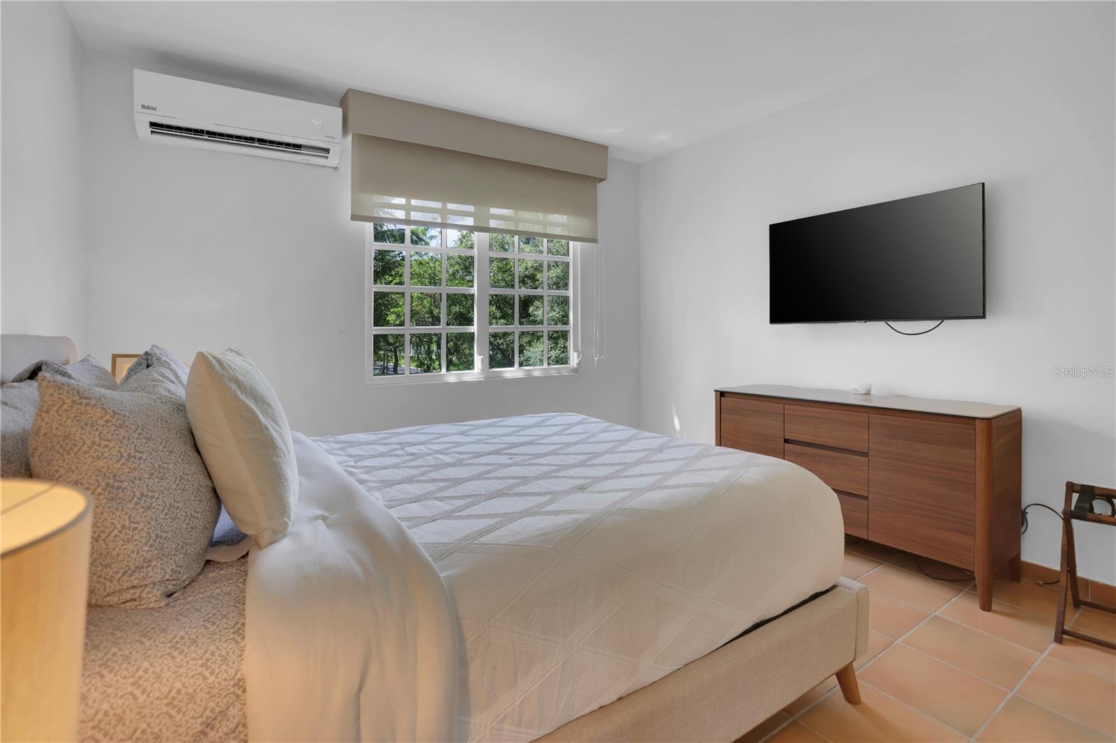 Bedroom with a large bed, leafy window view, and wall-mounted television above a wooden dresser.