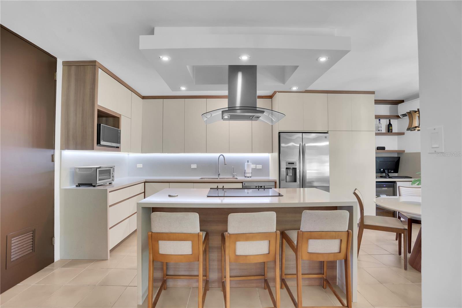Modern kitchen with a large island, three wooden chairs, cream cabinets, and stainless-steel appliances.