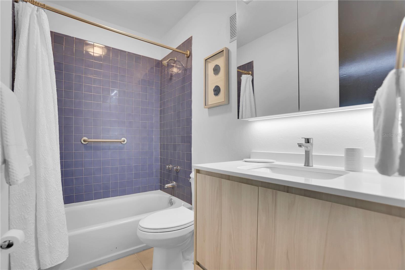 Bathroom with a purple-tiled tub and shower, white toilet, and light wood vanity.