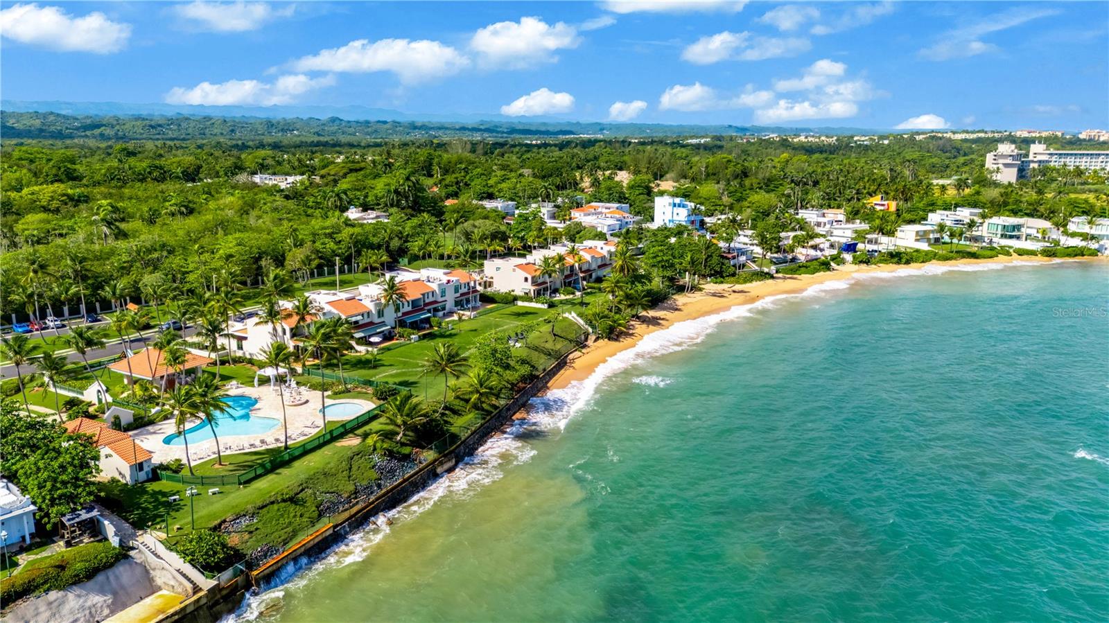 Aerial view of coastal homes beside turquoise water, a sandy beach, palm trees, and a swimming pool.