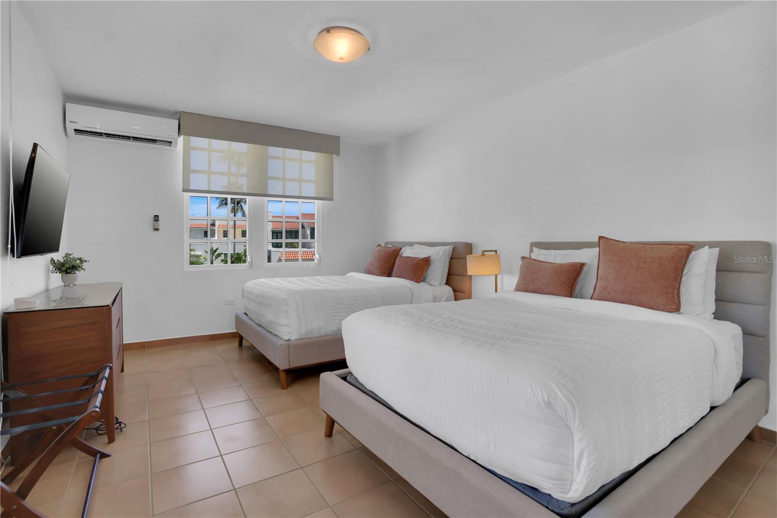 Bedroom with two white beds, rust-colored pillows, tiled floors, and a wall-mounted air conditioner.