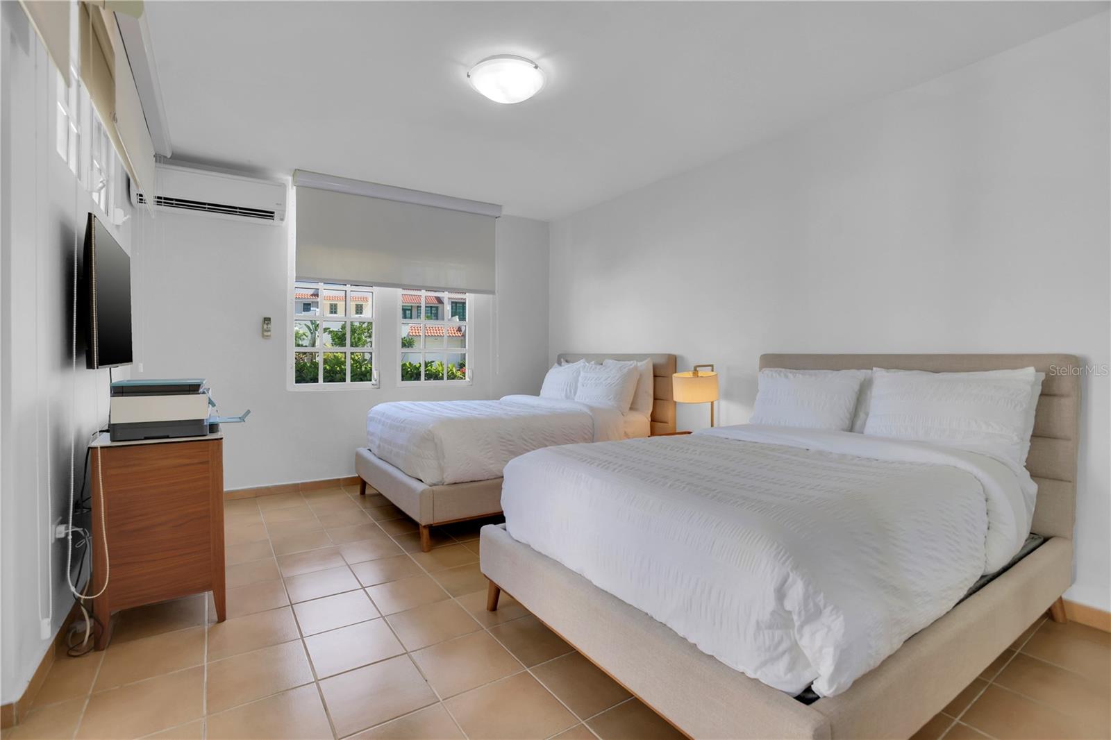 Bedroom with two white beds, tiled floors, and windows overlooking neighboring buildings.