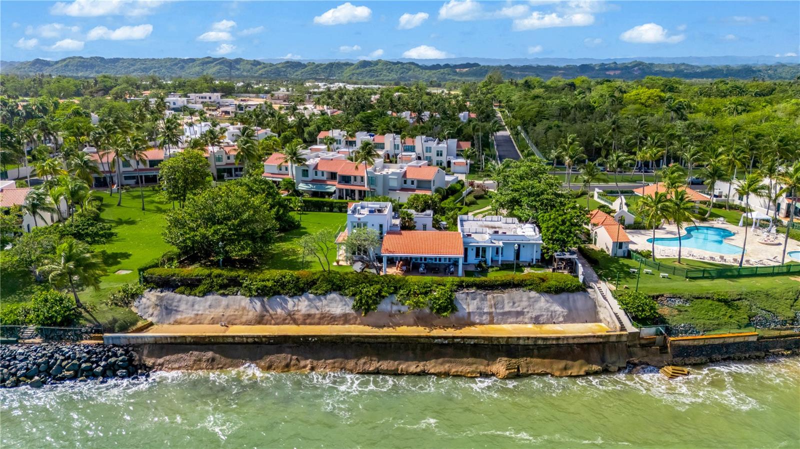 Aerial view of a waterfront home with a seawall, palm trees, and a neighboring pool.