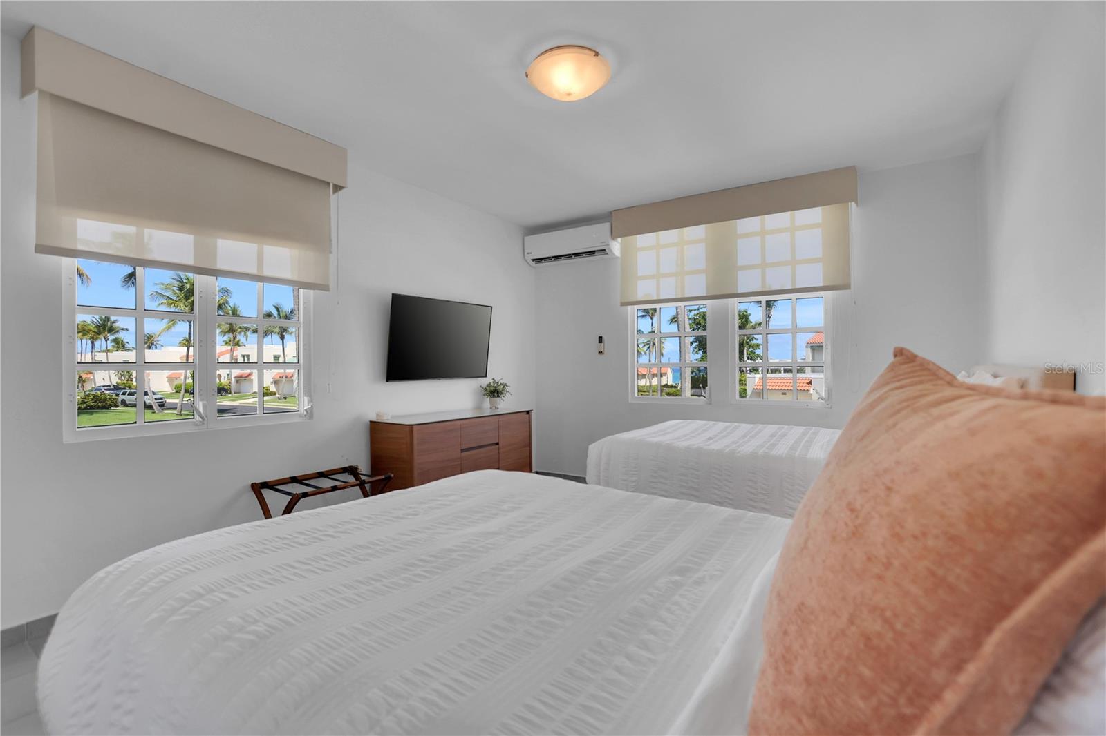 Bedroom with two white-covered beds and large windows overlooking palm trees and nearby homes.
