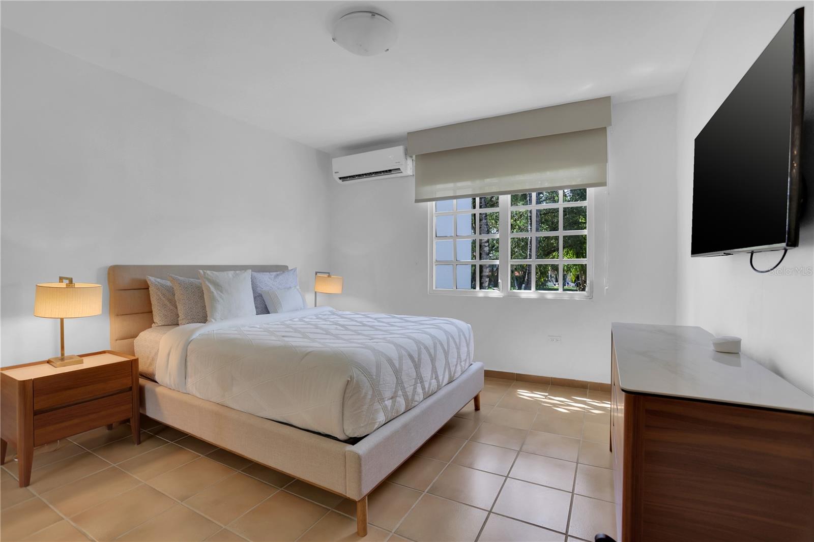 Furnished bedroom with a beige upholstered bed, tiled floor, gridded window, and wall-mounted television.