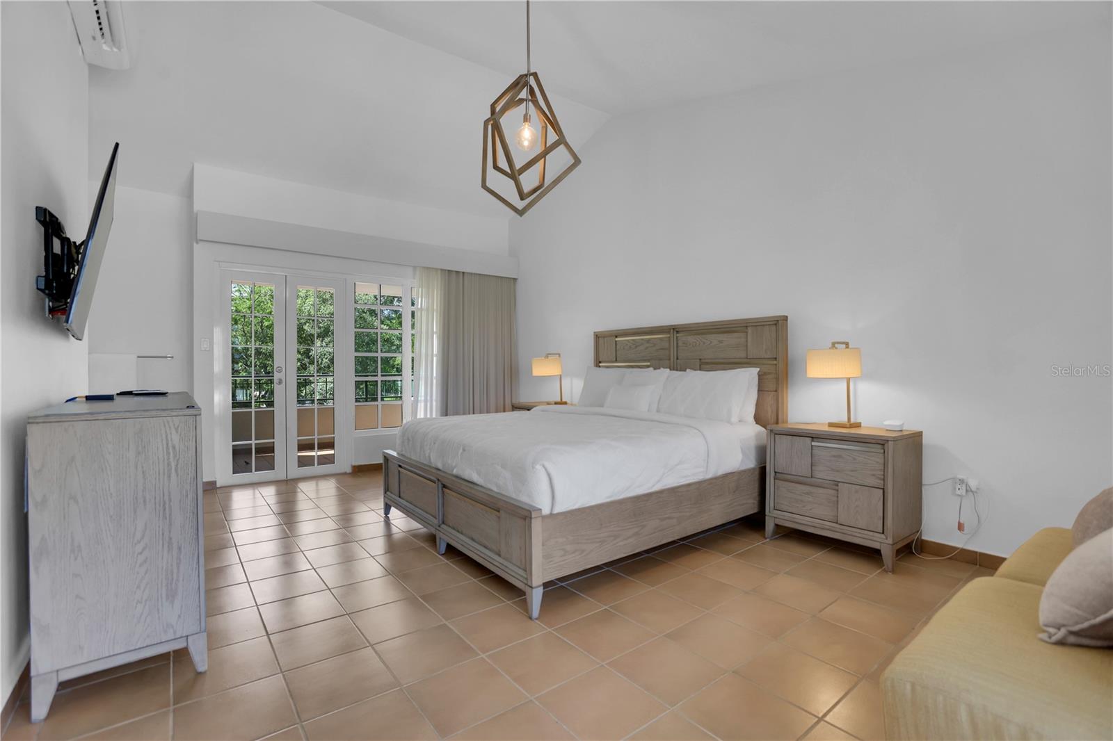 Furnished bedroom with a wooden bed, terracotta tile floor, and French doors overlooking greenery.