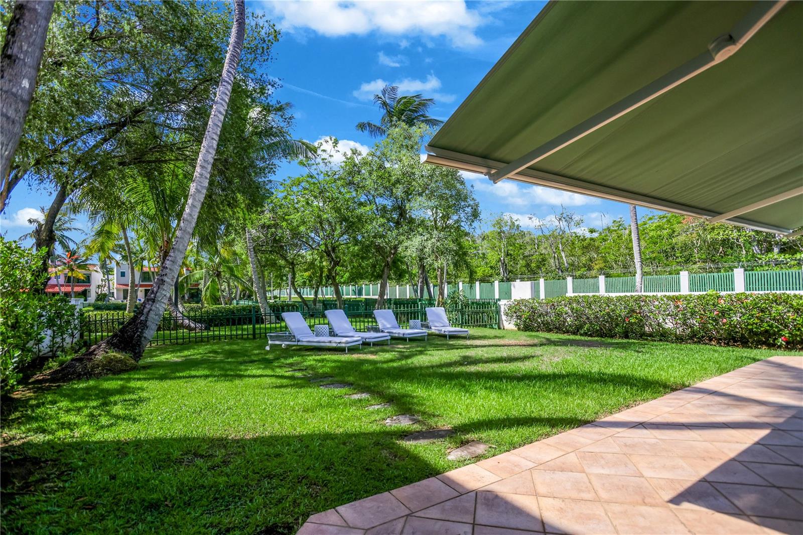 Backyard patio with a green awning, tropical trees, lawn, and four white lounge chairs.
