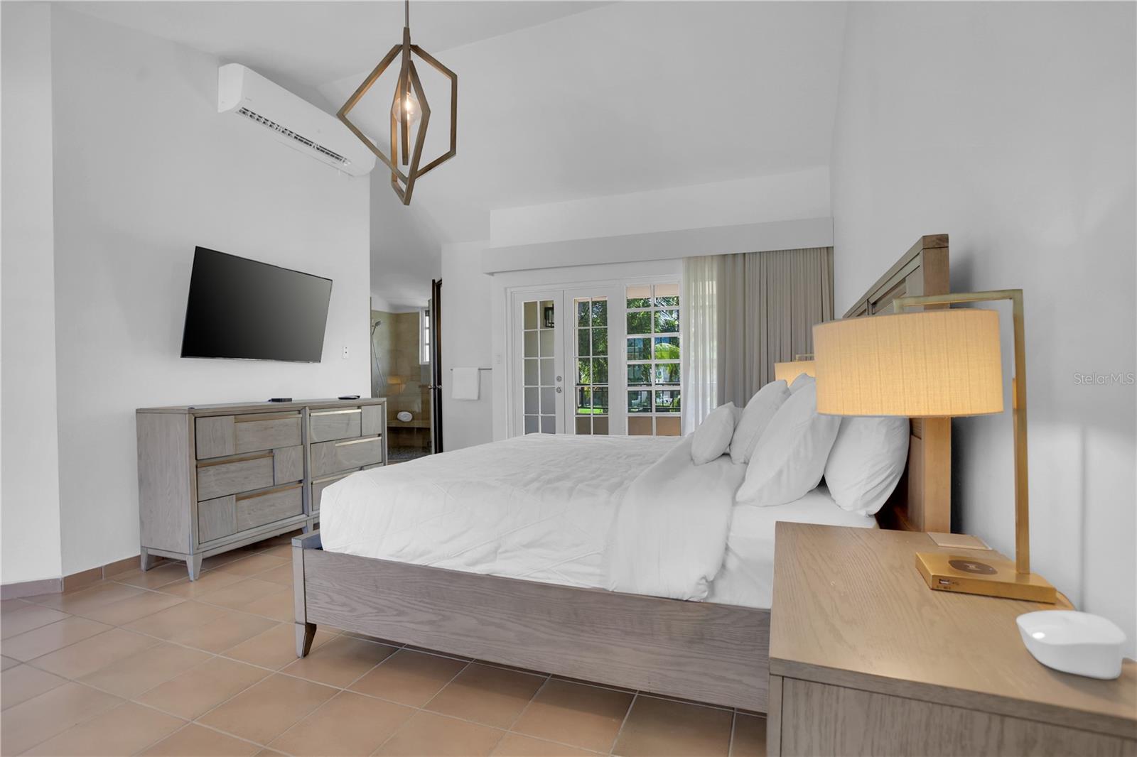 Furnished bedroom with a white bed, wood nightstand, and French doors overlooking greenery.