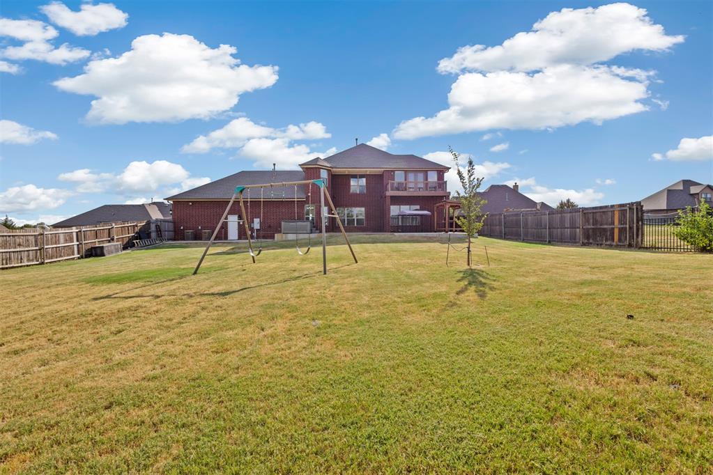 Fenced backyard with a broad lawn, wooden swing set, and two-story brick house.