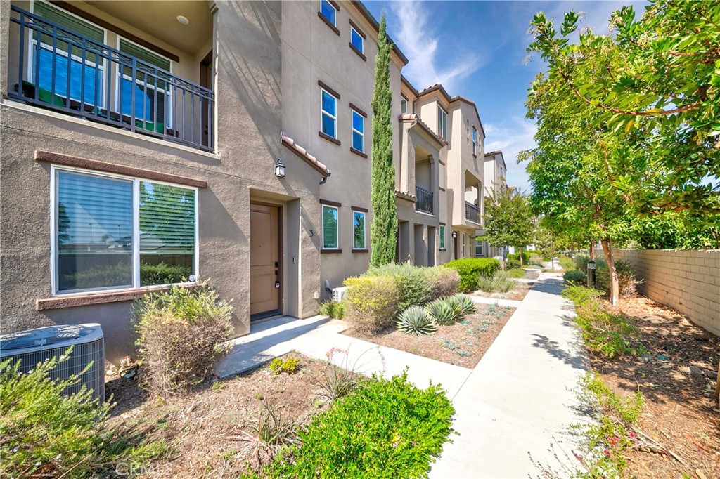 Attached stucco homes with balconies line a landscaped sidewalk beneath a mature leafy tree.