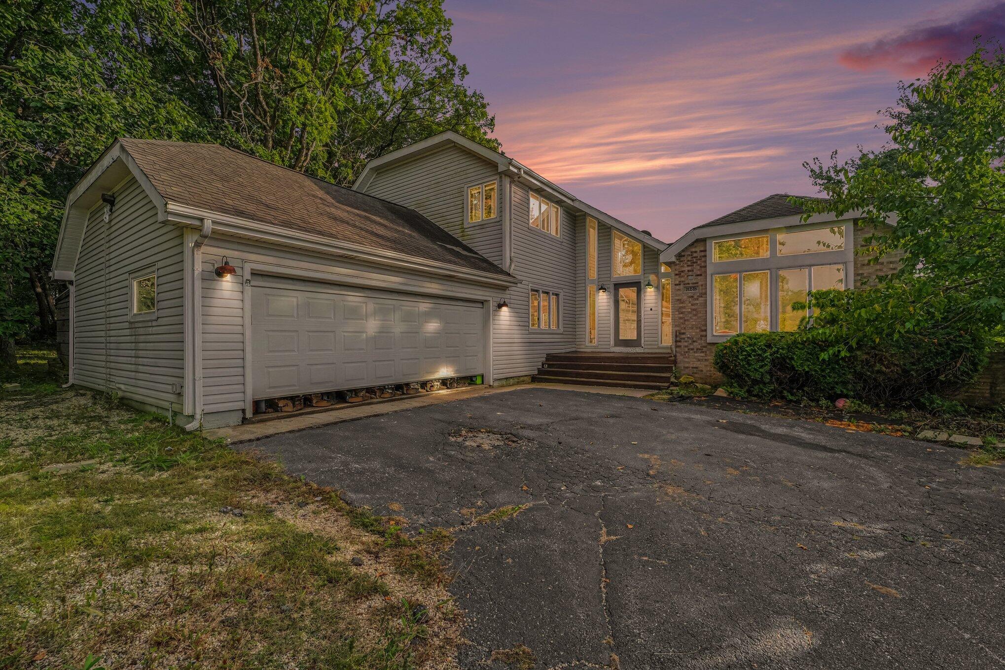 Gray multi-level home with a two-car garage and broad windows, surrounded by trees at dusk.