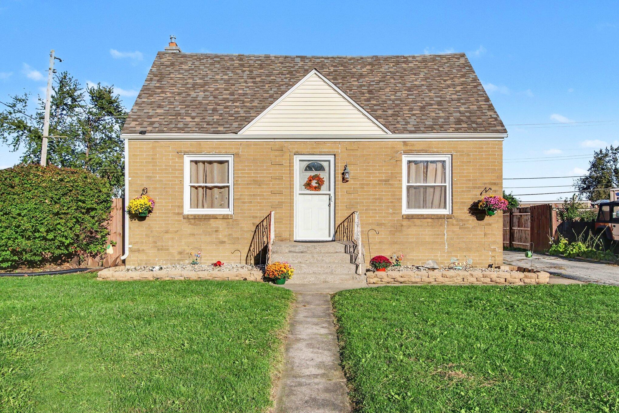 Tan brick house with a steep roof, flower beds, and a walkway across the lawn.