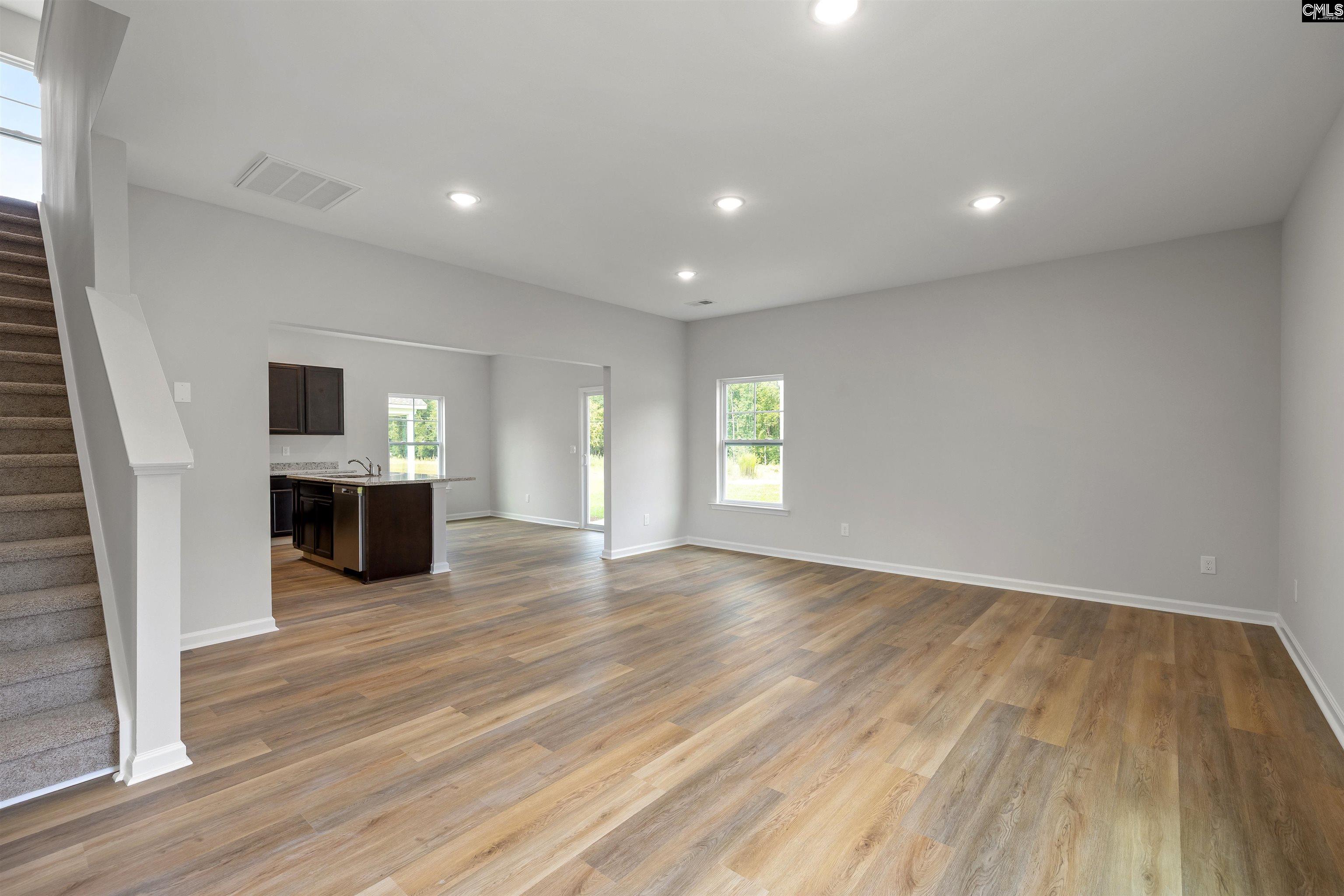 Open-plan living room with wood-look flooring, dark kitchen island, and carpeted stairs.