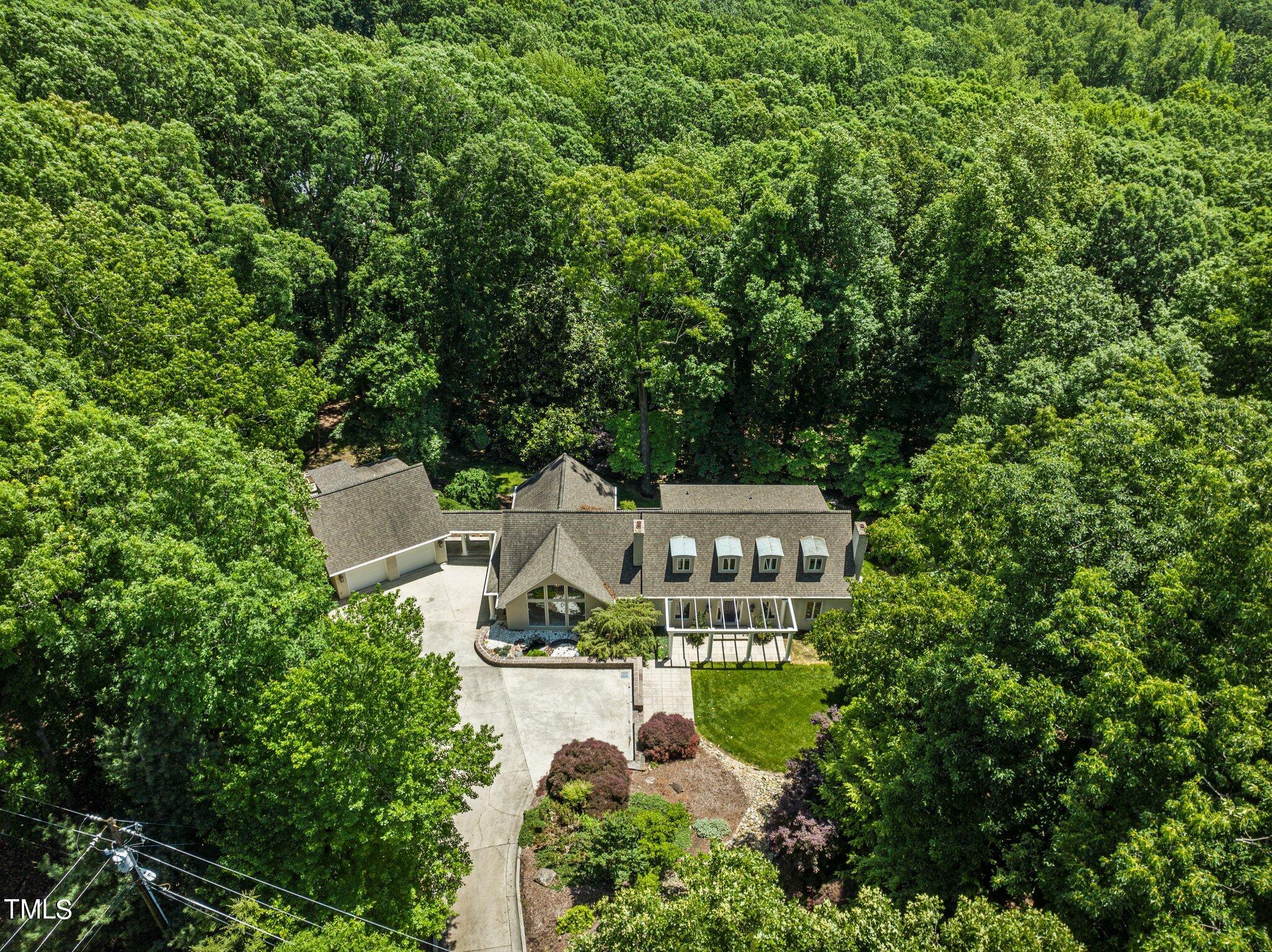Aerial view of a wooded home with dormer windows, an attached garage, and a broad driveway.