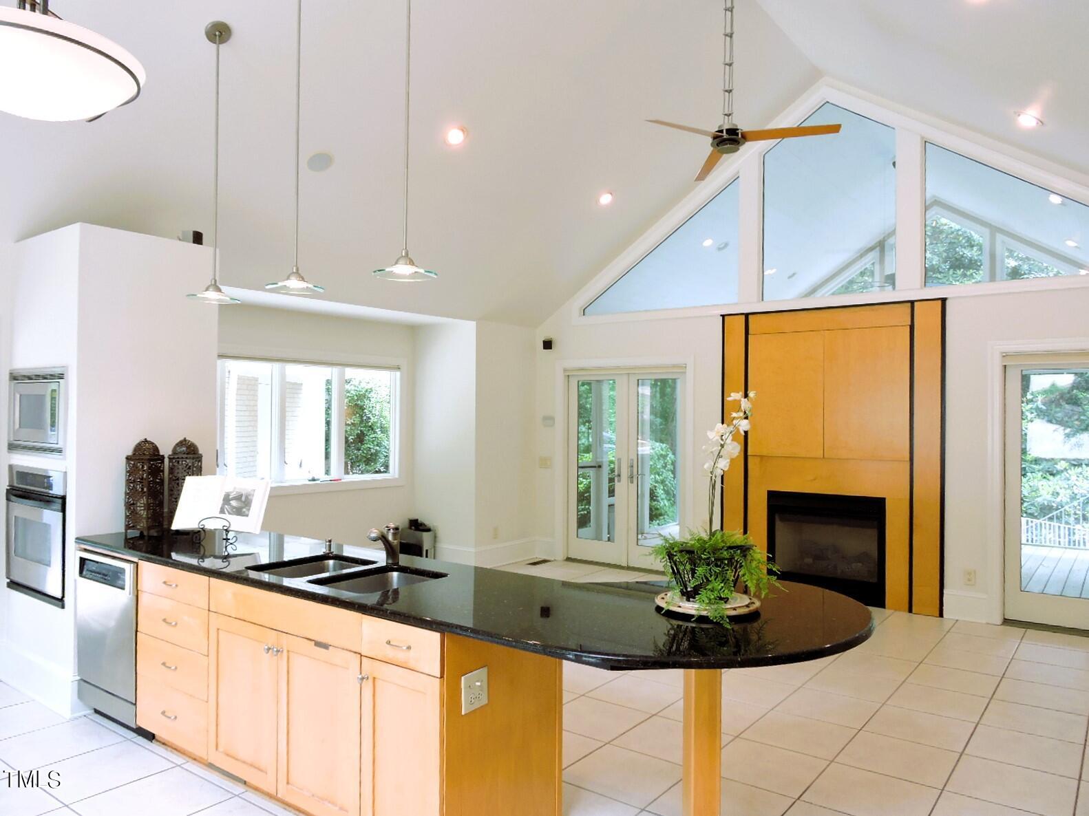 Open kitchen with a black granite island, light wood cabinets, and a fireplace beneath a vaulted ceiling.