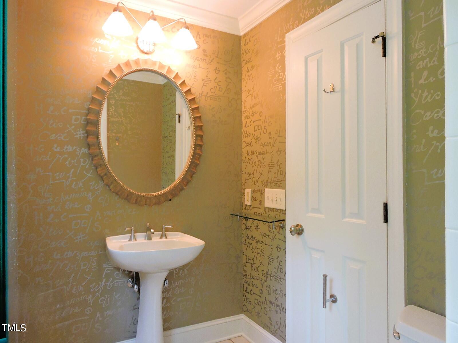 Bathroom with a pedestal sink, oval sunburst mirror, and script-patterned wallpaper.