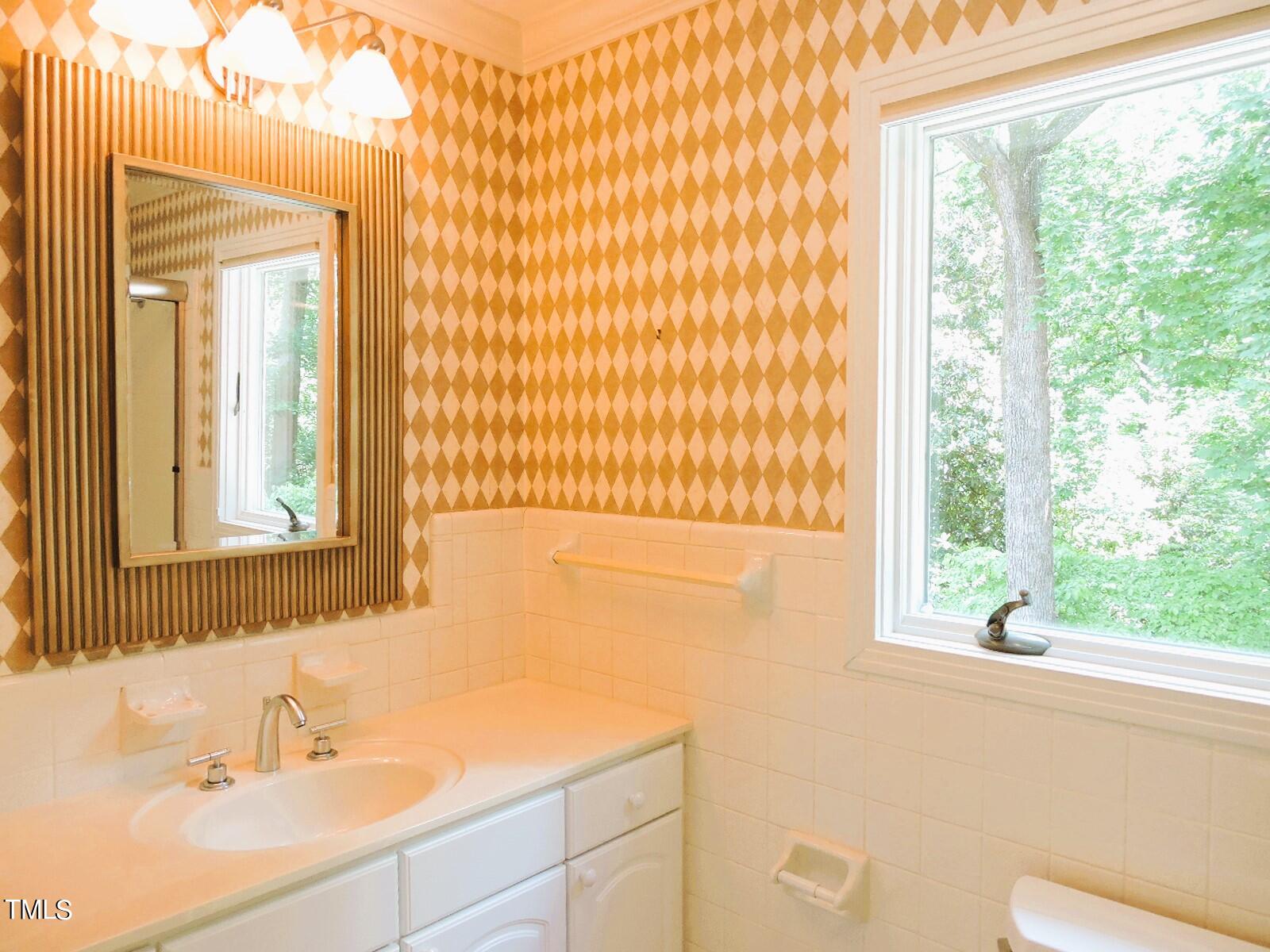 Bathroom vanity and mirror beside diamond-patterned wallpaper and a large window overlooking trees.